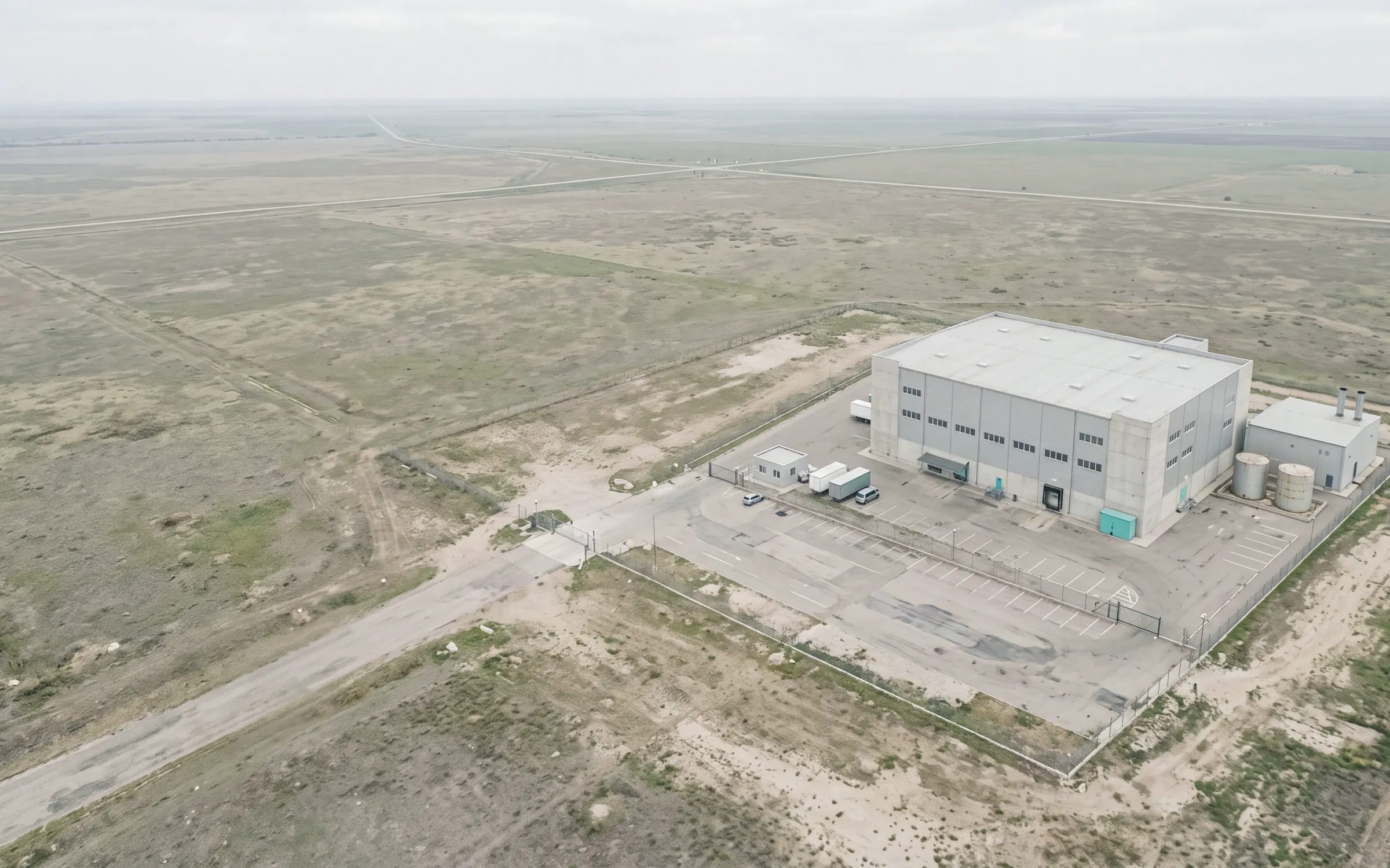 AI-generated scene: a logistics park with its long warehouse, tanks and fenced yard, seen from the air.